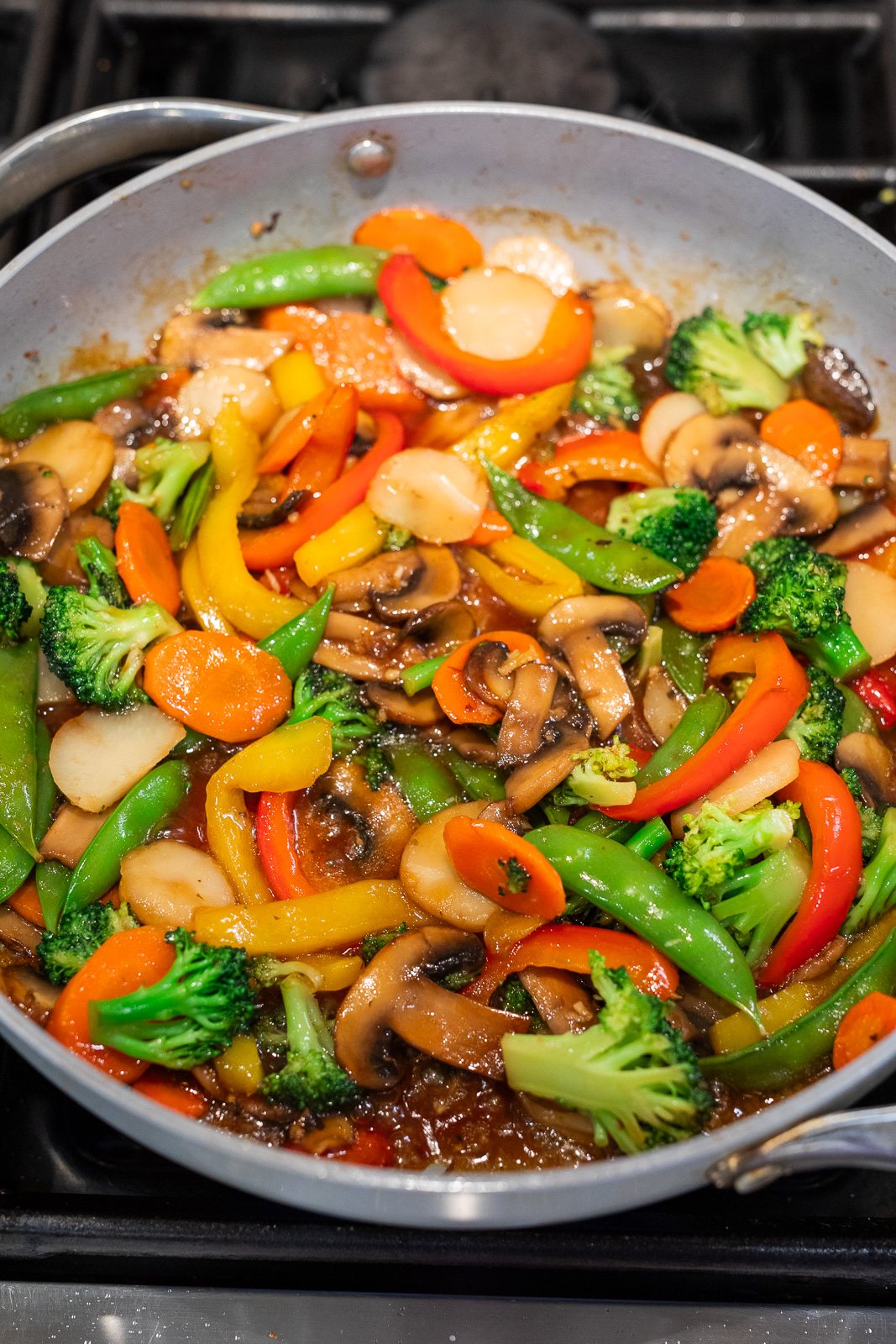 simple vegetable stir fry simmering in a skillet with glossy stir fry sauce coating broccoli, mushrooms, bell peppers, carrots, and snap peas
