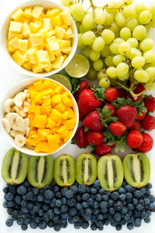 Fresh fruit salad ingredients including mango, pineapple, strawberries, kiwi, bananas, blueberries, and grapes on a table