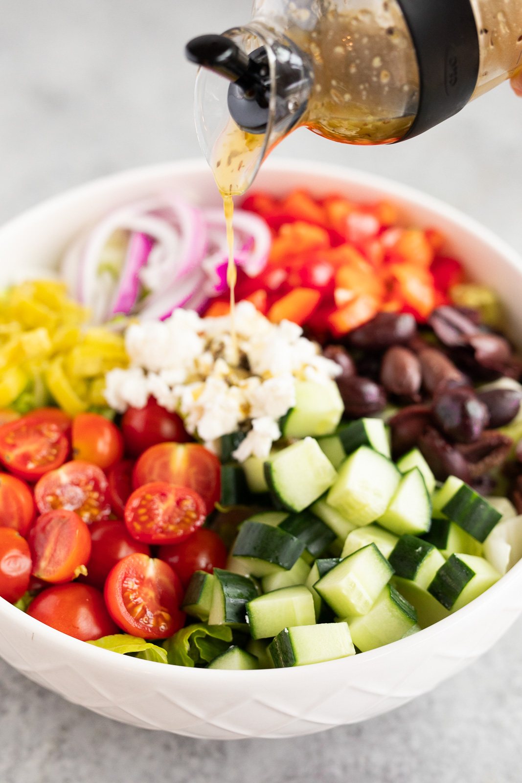 Pouring homemade Italian dressing over chopped salad with romaine, cucumbers, tomatoes, bell pepper, red onion, olives, and feta