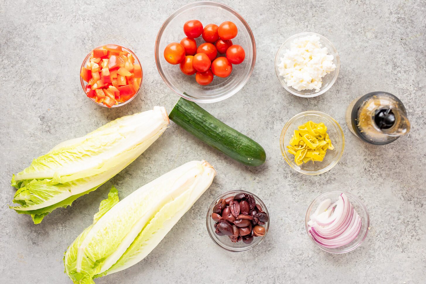 Ingredients for easy chopped salad including romaine lettuce, cherry tomatoes, red bell pepper, cucumber, red onion, olives, feta, pepperoncini, and homemade Italian dressing