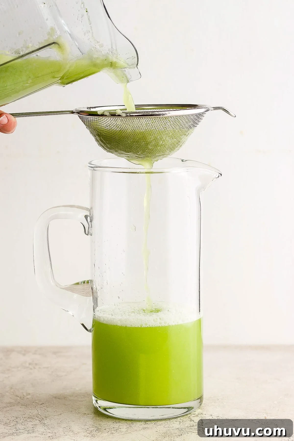Agua de Pepino mixture being carefully strained through a fine-mesh sieve into a large, clear glass pitcher to achieve a smooth consistency.
