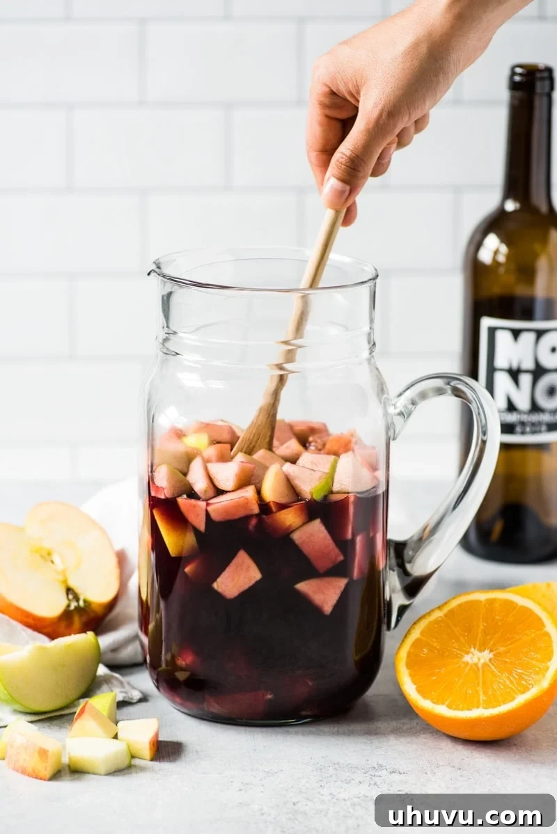 A large glass pitcher of red sangria is being gently mixed with a spoon, revealing an abundance of colorful fruit pieces.