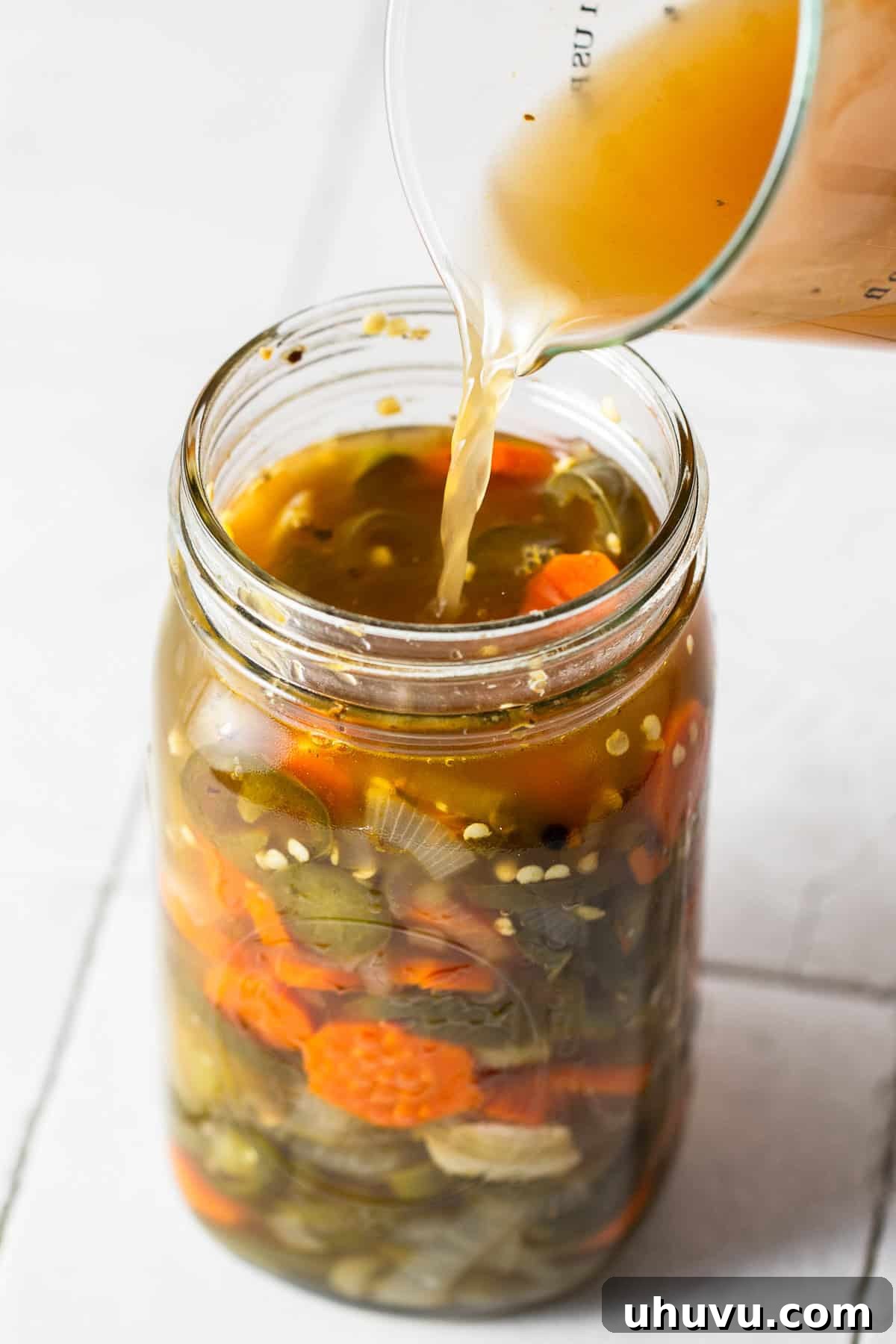 Zesty Pepper Bites 6 Pickling liquid being poured into a jar of pickled jalapenos. A clear, hot brine is carefully poured over the sautéed vegetables, ensuring they are fully submerged for optimal pickling.