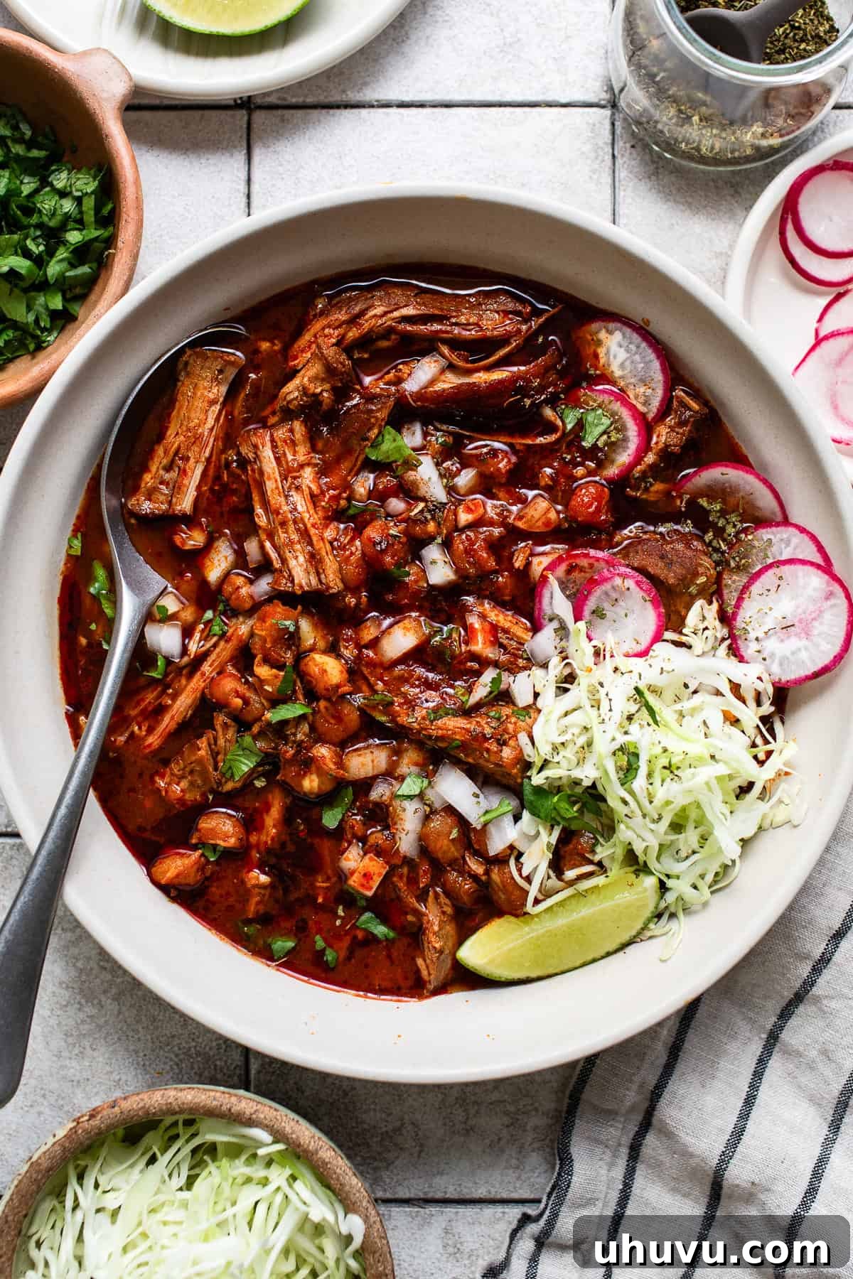 A large bowl of vibrant red pozole rojo, garnished with fresh lime wedges, a sprinkle of Mexican oregano, thinly sliced radishes, aromatic cilantro, and crisp shredded cabbage, ready to be enjoyed.