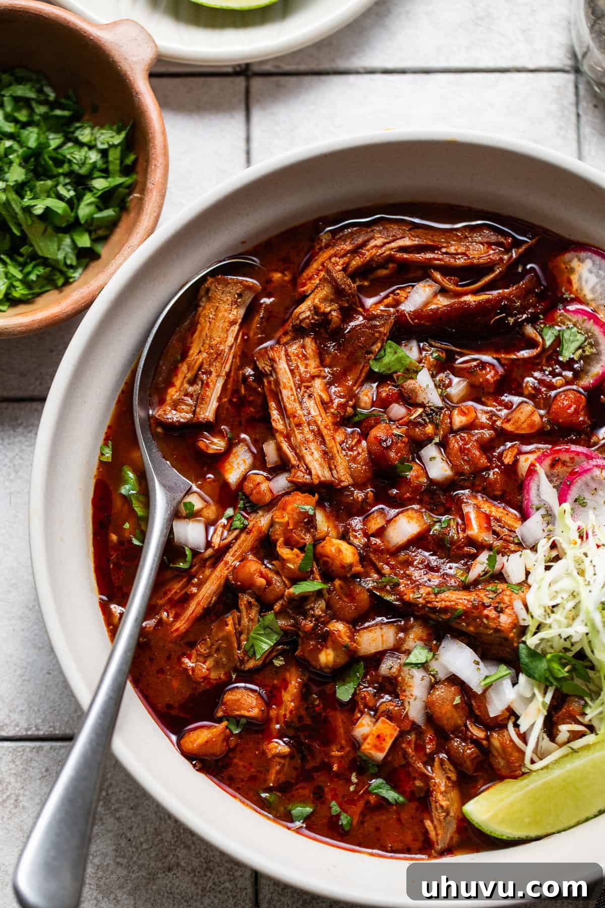 A steaming bowl of delicious Mexican pozole rojo, generously topped with crisp shredded cabbage, bright green cilantro, zesty lime wedges, and crunchy radish slices, with diced onions peeking through.
