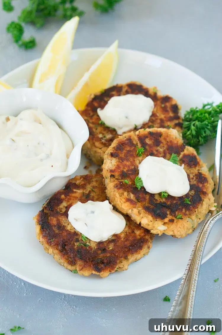 Two perfectly cooked salmon patties served on a white plate, elegantly topped with creamy tartar sauce and a sprinkle of fresh herbs.