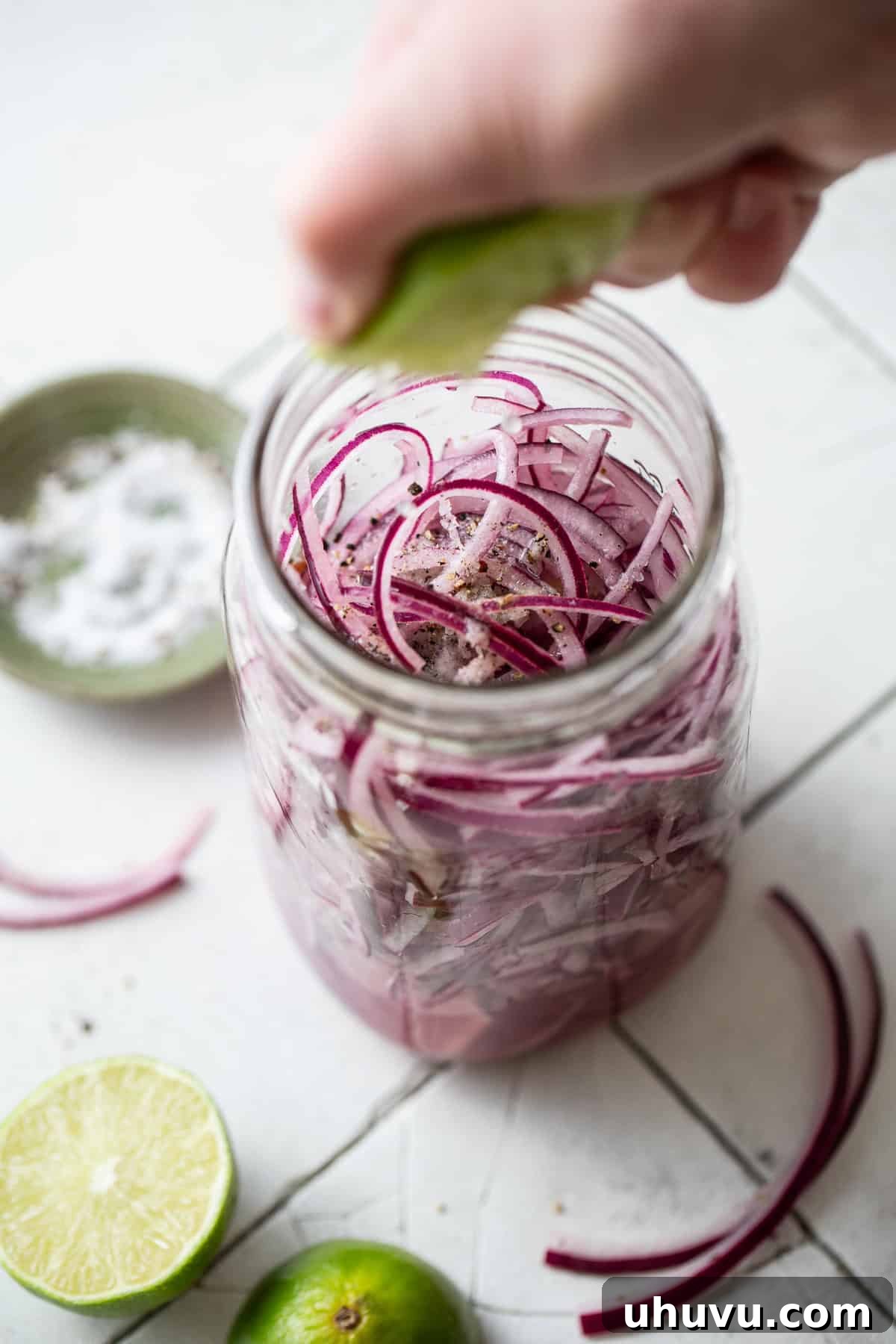 Fresh lime juice being squeezed directly into a glass jar filled with thinly sliced red onions, seasoned with kosher salt and black pepper.