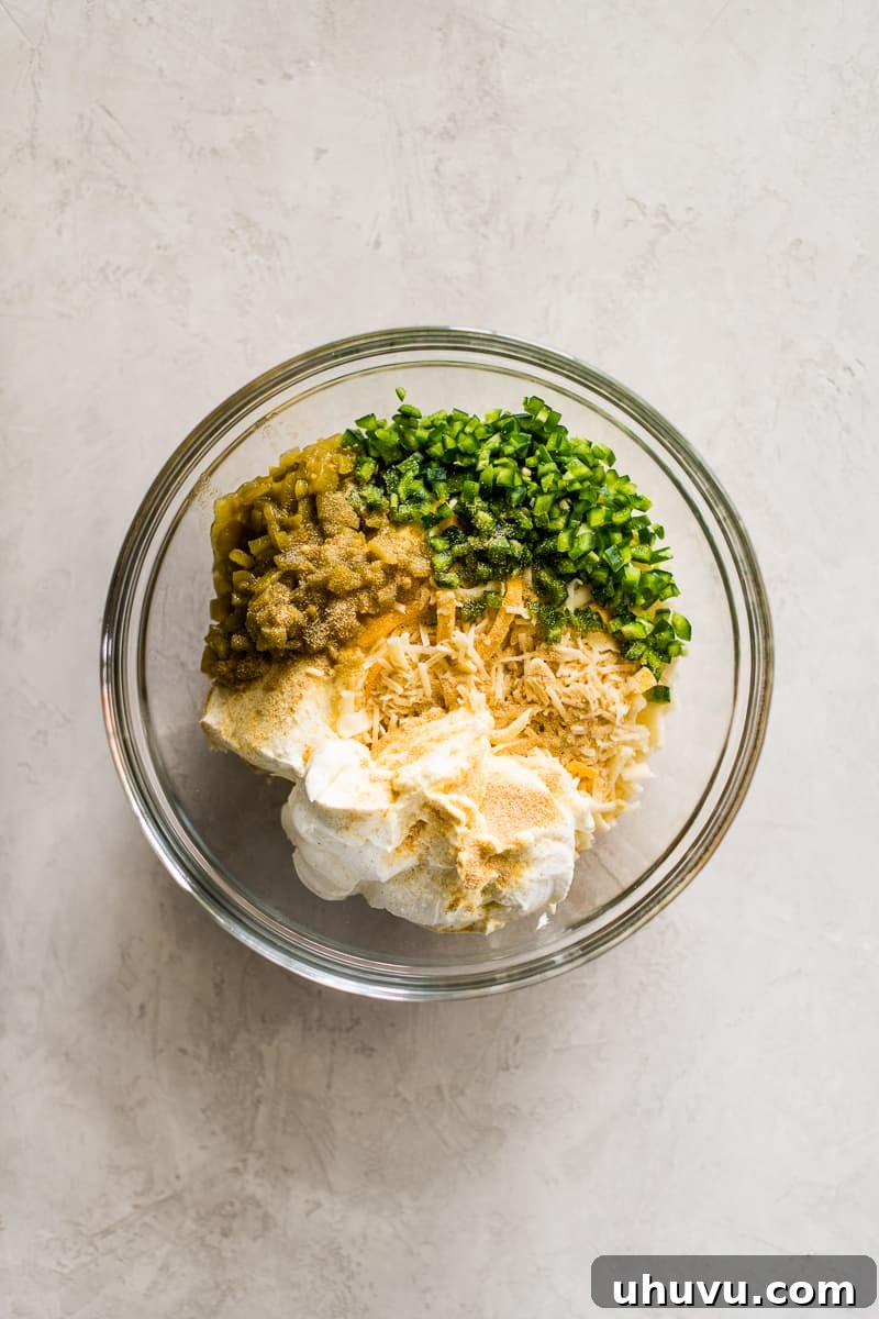 A mixing bowl filled with cream cheese, sour cream, shredded cheese, diced green chiles, jalapenos, and seasonings.
