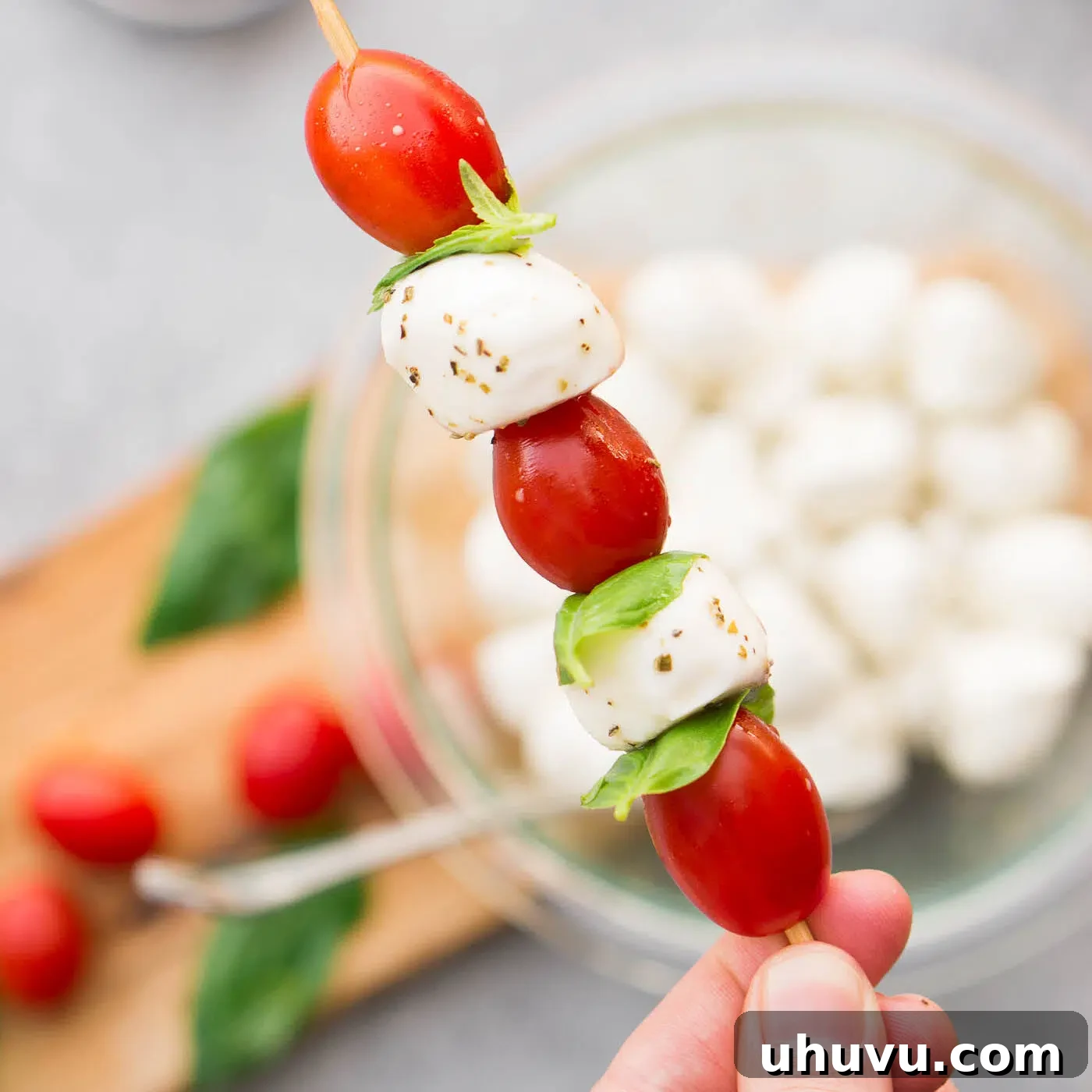 Hands carefully threading cherry tomatoes, basil leaves, and marinated mozzarella balls onto wooden skewers
