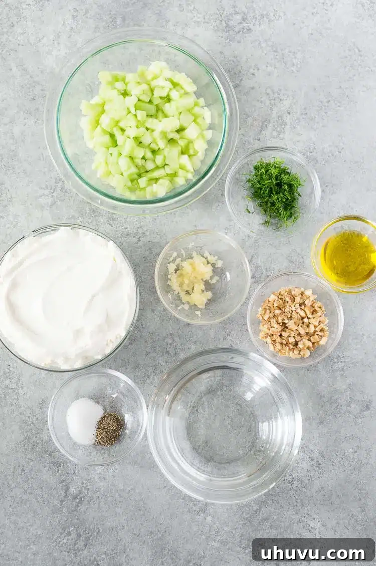 Fresh ingredients for Tarator: cucumber, garlic, dill, olive oil, salt, pepper, yogurt, water, and walnuts arranged on a gray board.