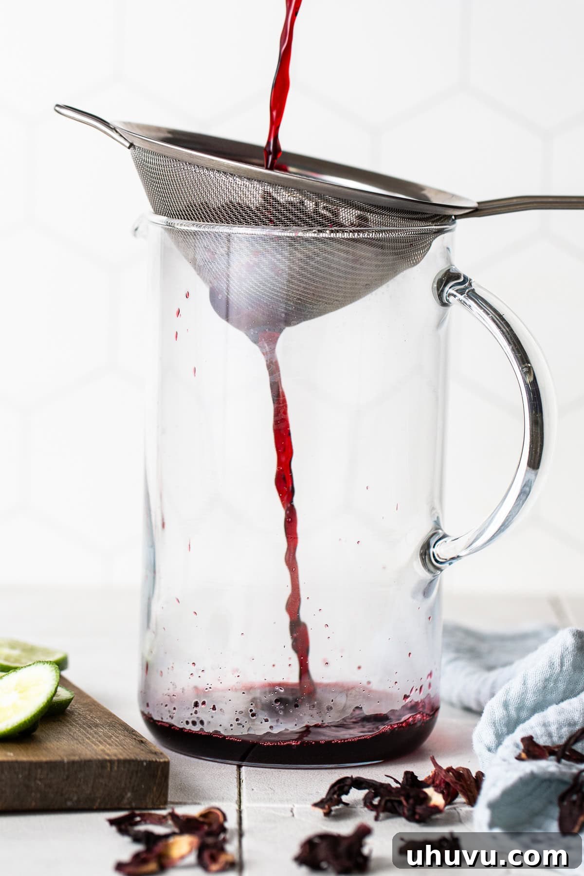 Ruby Hibiscus Refresher 5 Hot hibiscus tea being poured through a fine-mesh strainer into a large glass pitcher.