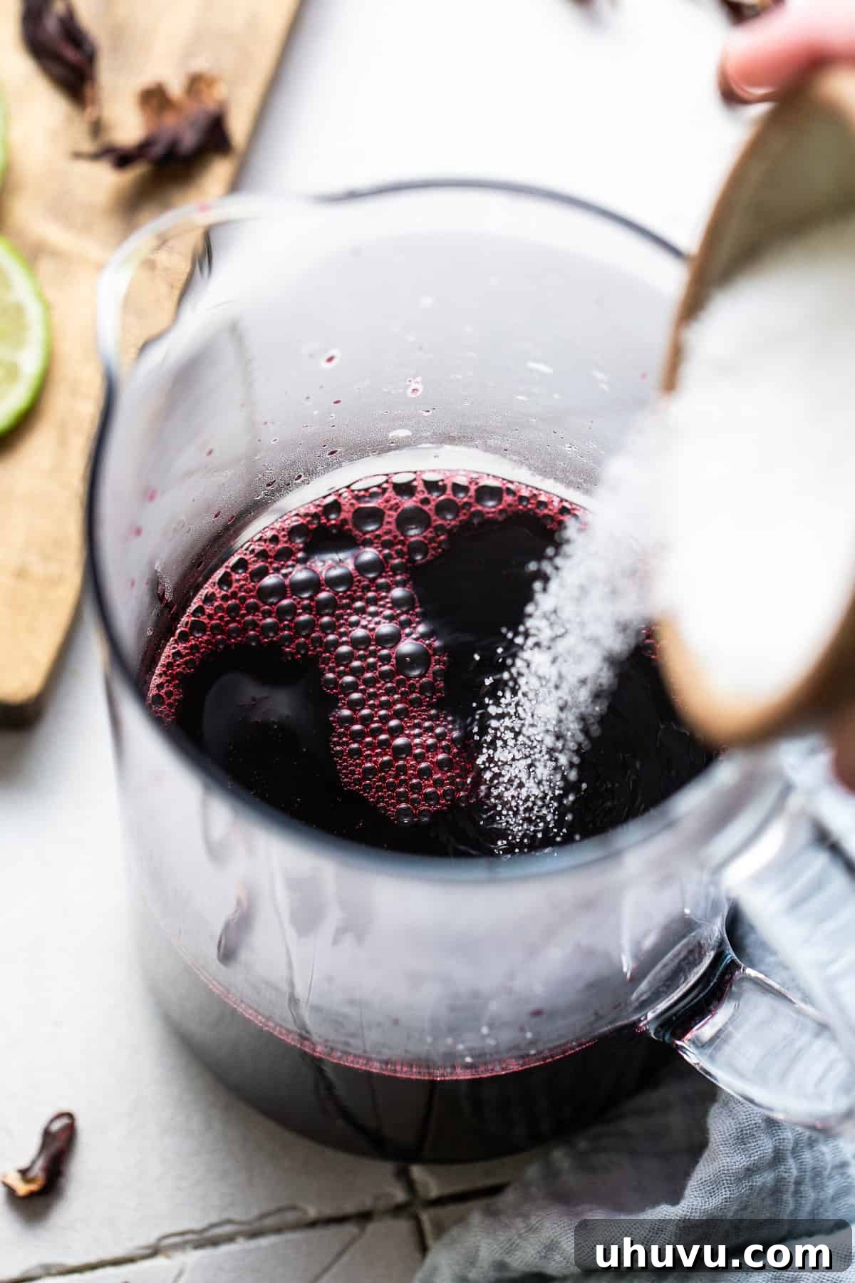 Ruby Hibiscus Refresher 6 Granulated sugar being poured into a pitcher of agua de jamaica, ready to be stirred.