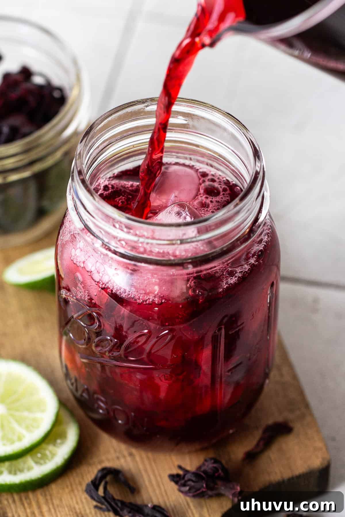 Ruby Hibiscus Refresher 7 Agua de Jamaica drink being poured from a pitcher into a tall glass filled with ice.