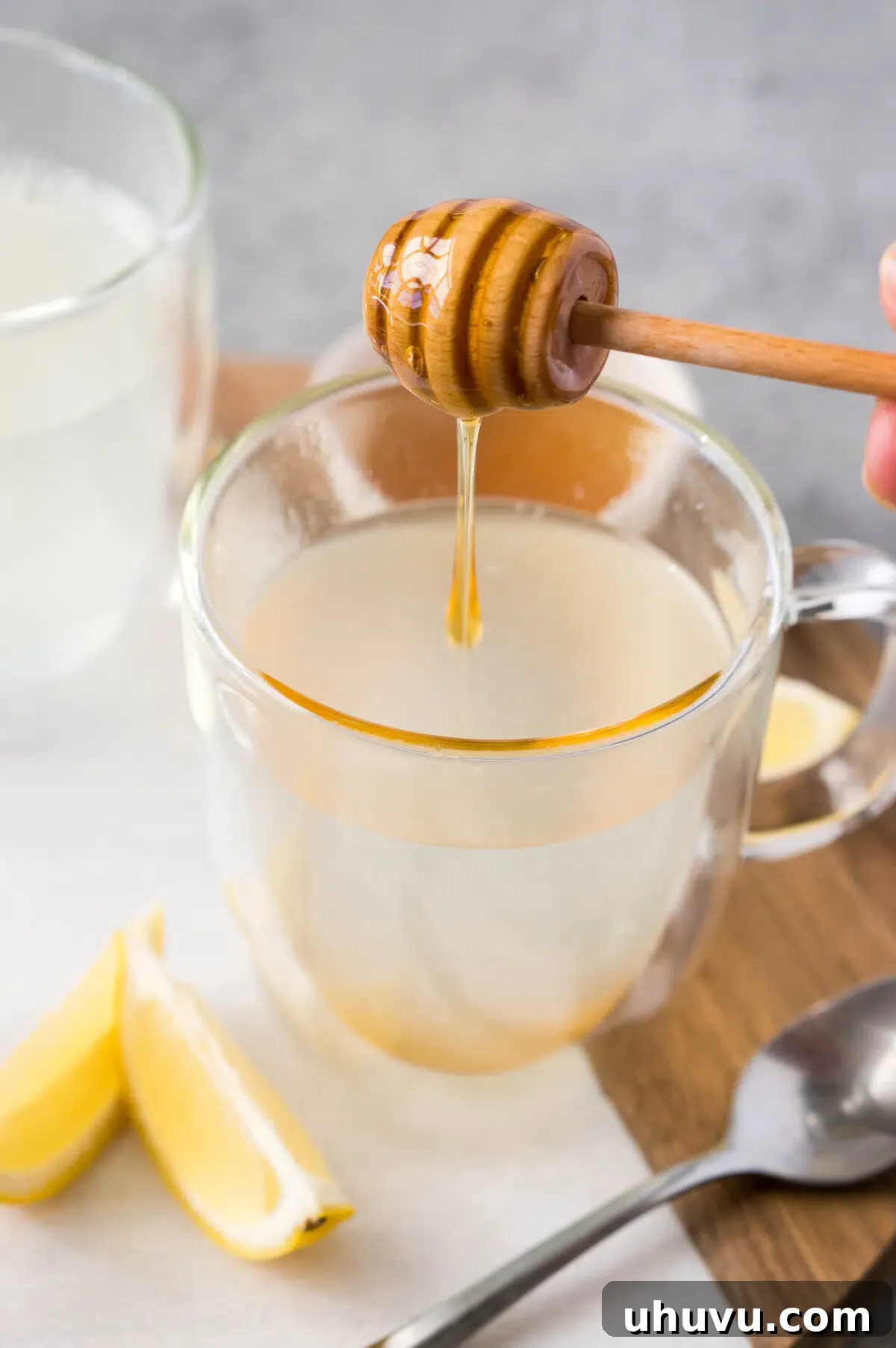 Adding a spoonful of honey to hot lemon tea in a clear glass cup.