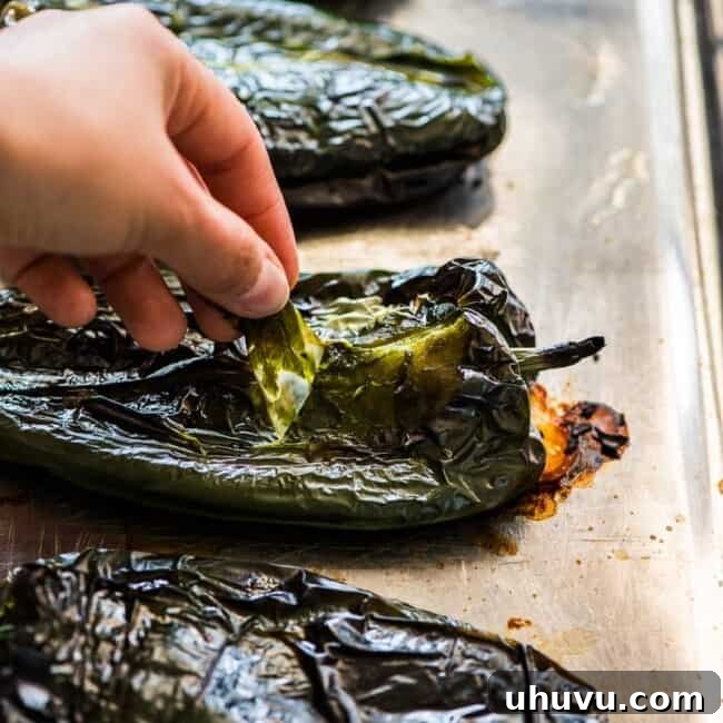 A hand pulling the skin off of roasted poblano peppers on a cutting board, with other roasted peppers in the background.