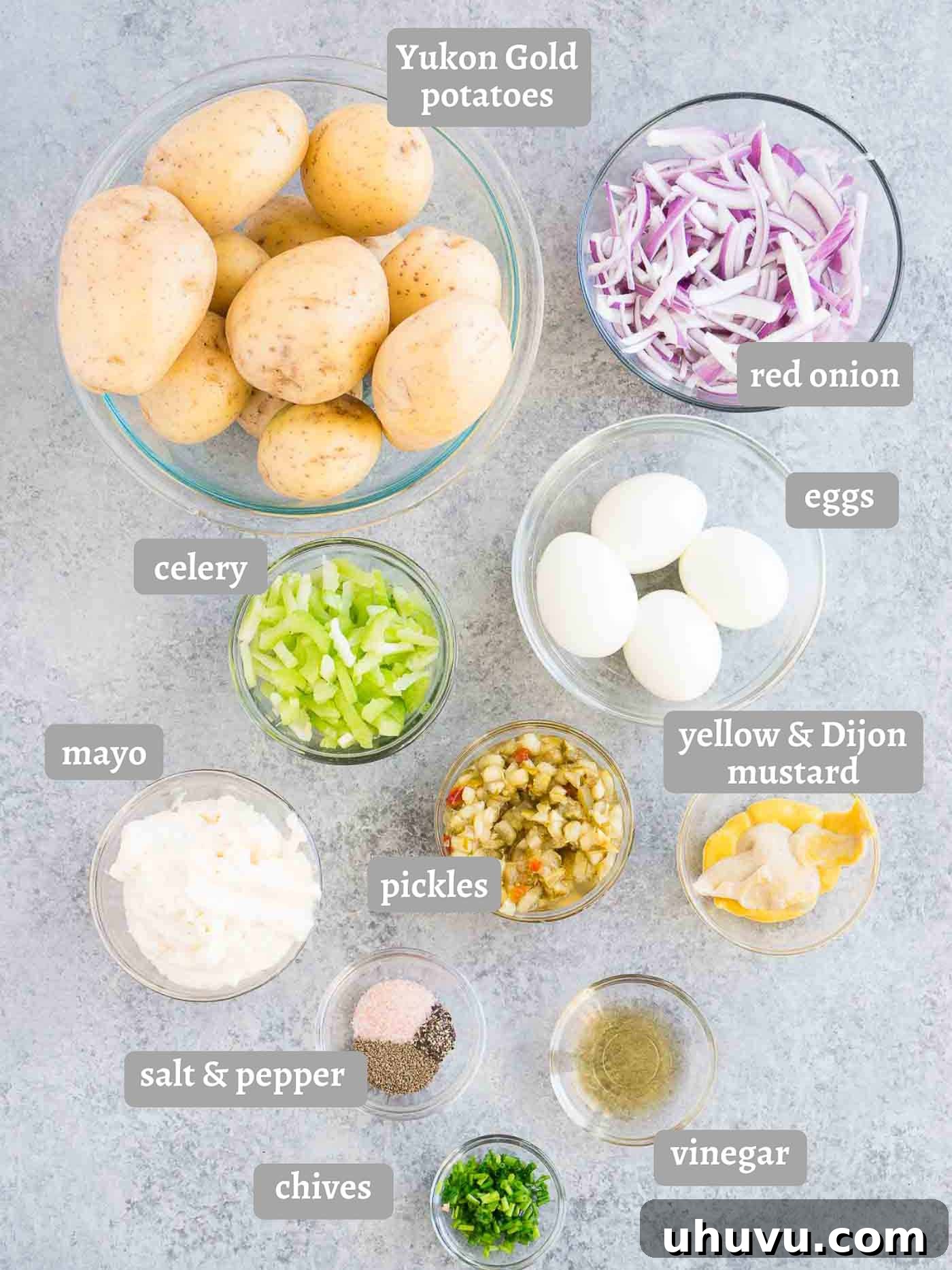 A selection of fresh potato salad ingredients laid out on a wooden surface, including Yukon Gold potatoes, celery, red onion, hard-boiled eggs, and various dressing components.