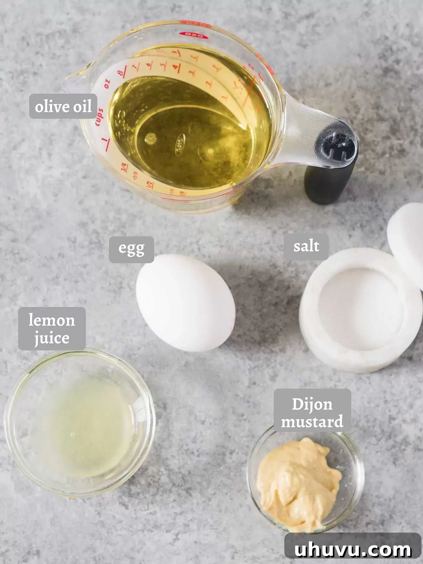 A flat lay photograph showcasing all the simple ingredients for homemade mayonnaise: an egg, olive oil, Dijon mustard, salt, and a lemon