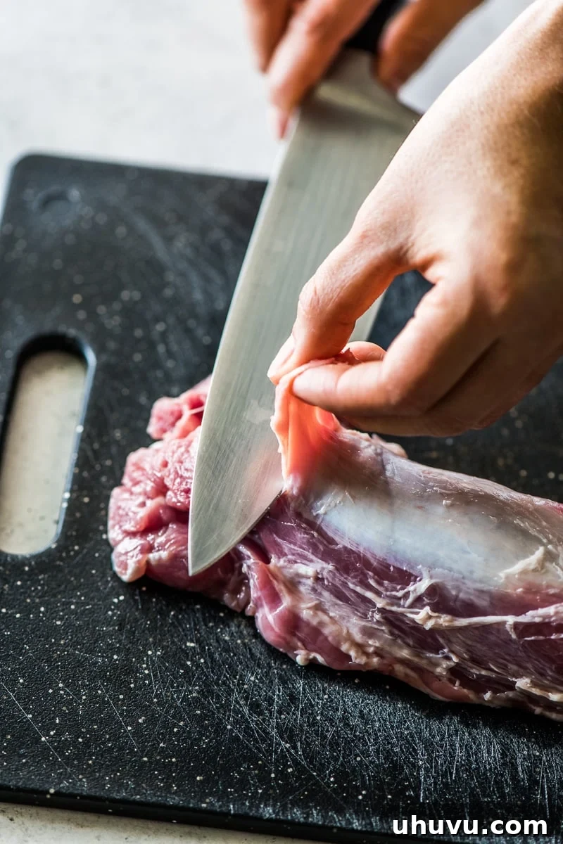 A knife and hand removing silver skin from a pork tenderloin.