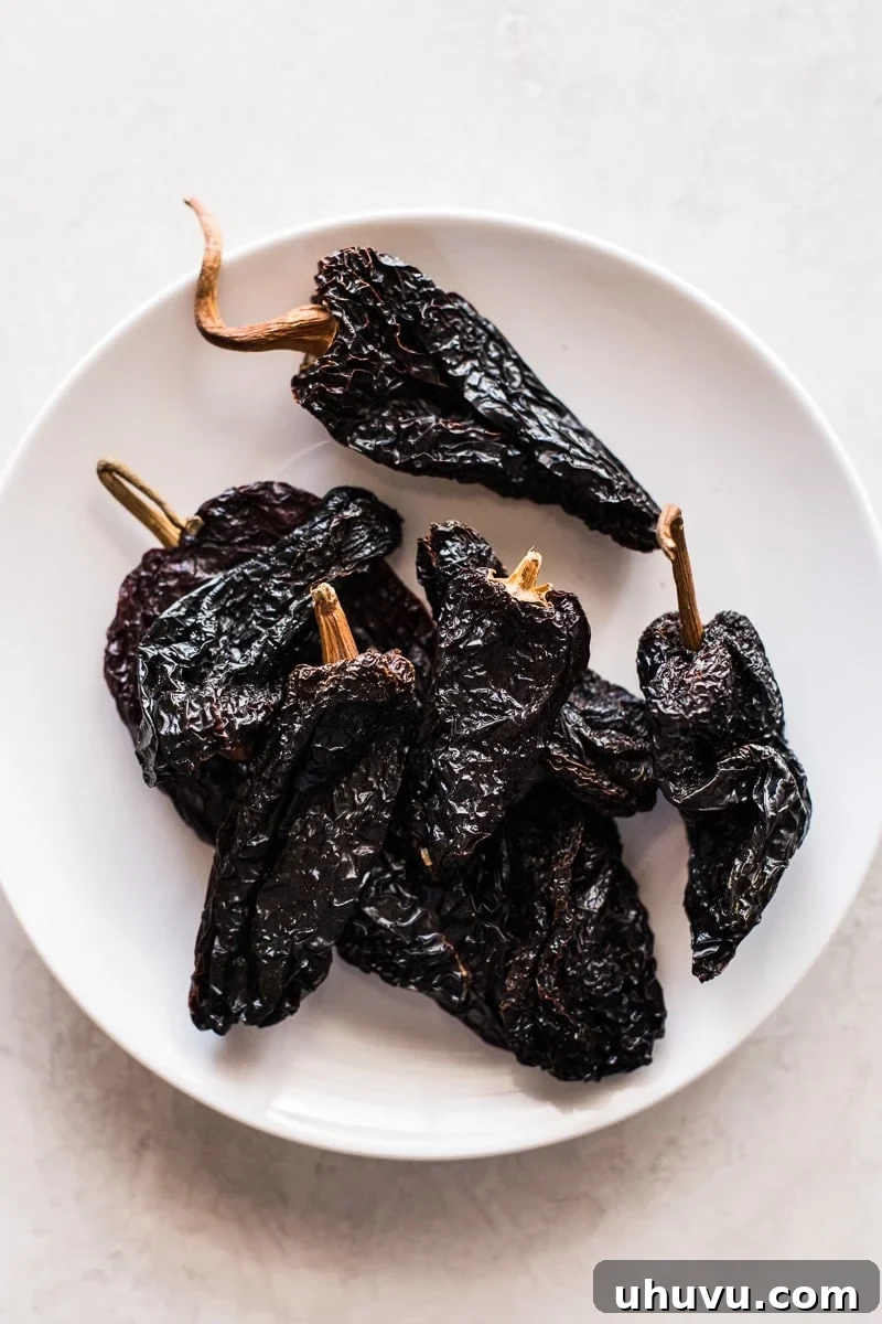 Smoky Sweet Anchos 2 Beautifully dried ancho chile peppers, with their dark burgundy color and leathery texture, resting on a pristine white plate, showcasing their culinary potential.