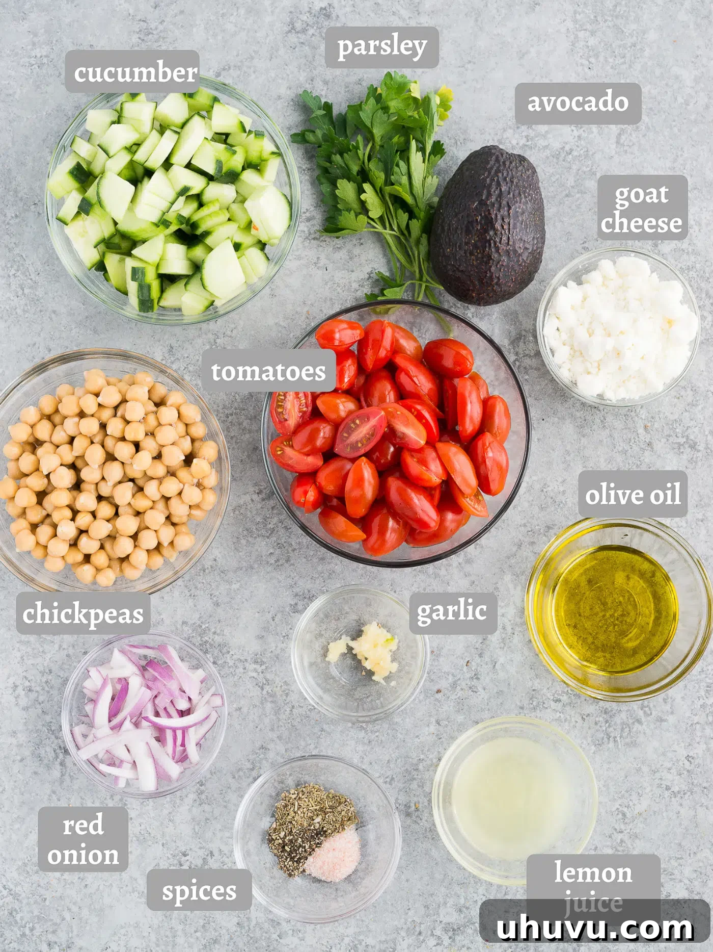 A wooden cutting board displaying all fresh ingredients for Mediterranean chickpea salad: cucumber, cherry tomatoes, red onion, chickpeas, avocado, goat cheese, and parsley.