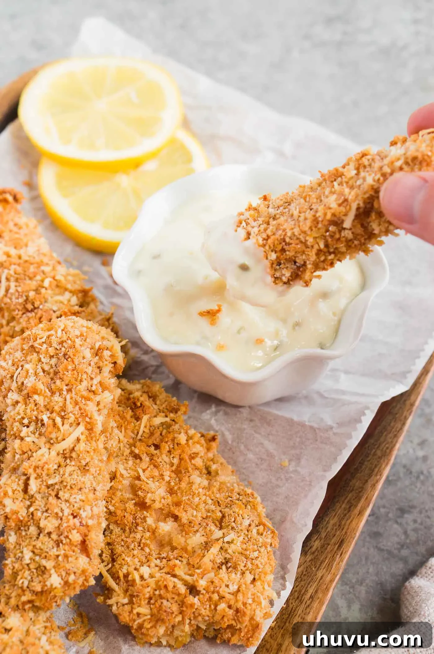 A close-up shot of a crispy homemade fish stick being dipped into a creamy white tartar sauce, highlighting its golden texture.