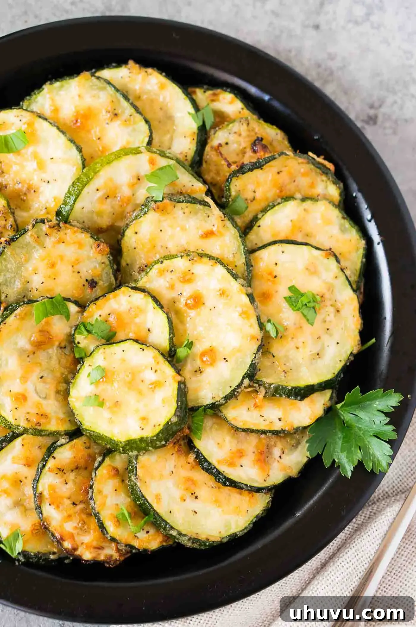Close-up view of oven-roasted zucchini slices on a white serving plate, showcasing their tender texture and golden-brown edges with melted Parmesan.