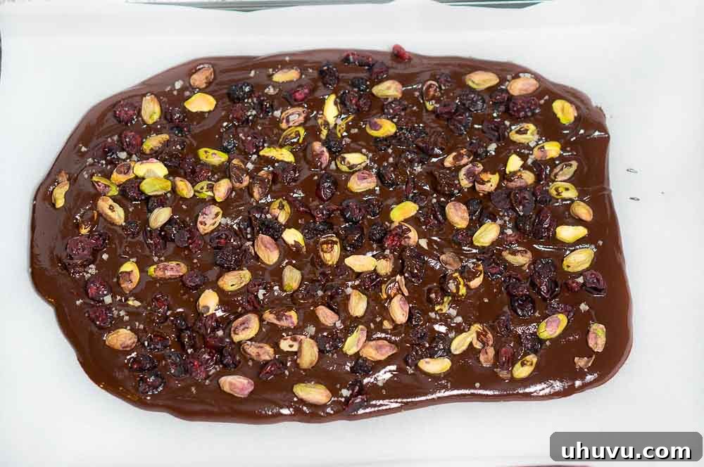 Crimson Pistachio Chocolate Bark 4 Chocolate mixture being spread onto a parchment-lined baking sheet, with pistachios and cranberries sprinkled on top.
