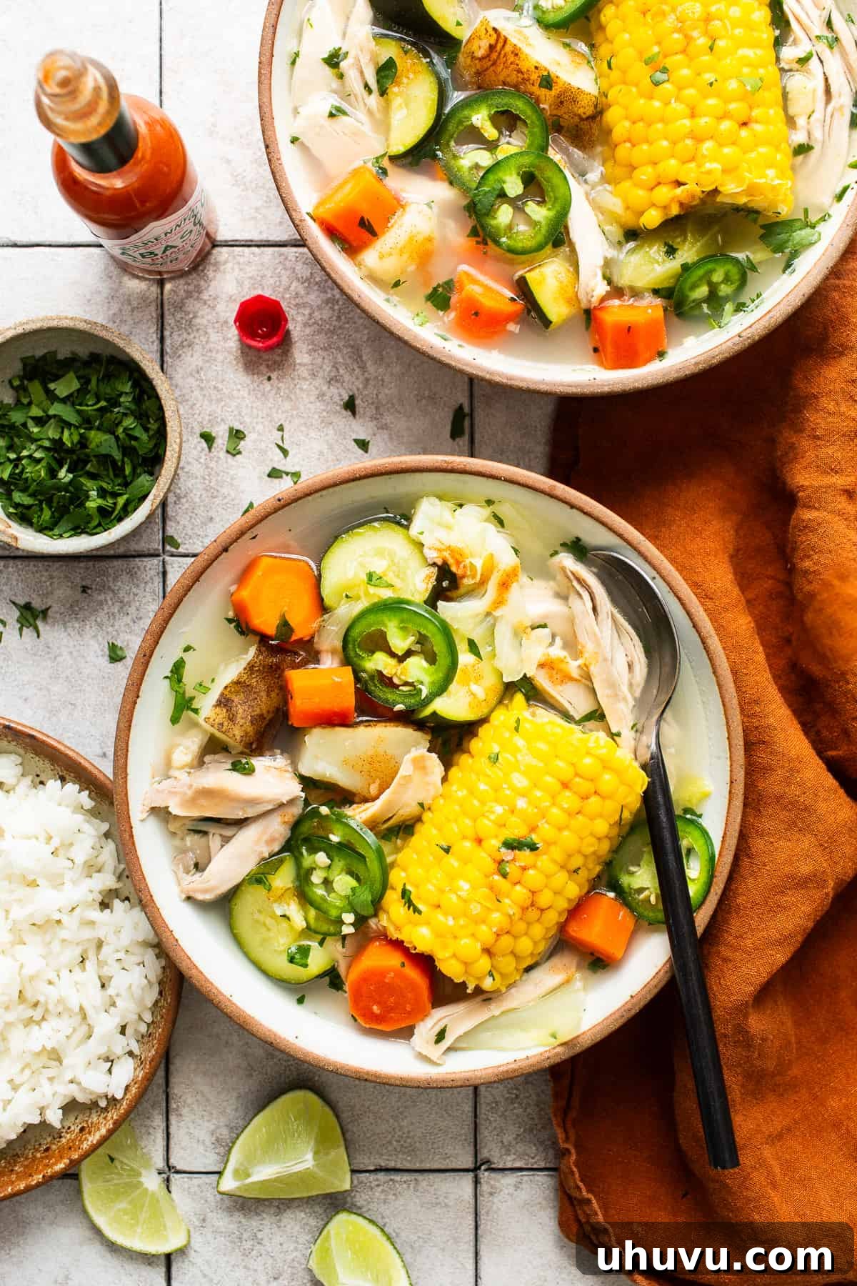 Wholesome Chicken Comfort 2 Two bowls of steaming Caldo de Pollo, Mexican chicken soup, garnished with fresh cilantro, alongside a serving of white rice, lime wedges, and a bottle of Tabasco sauce. The soup is rich with chicken, corn, carrots, potatoes, and zucchini.