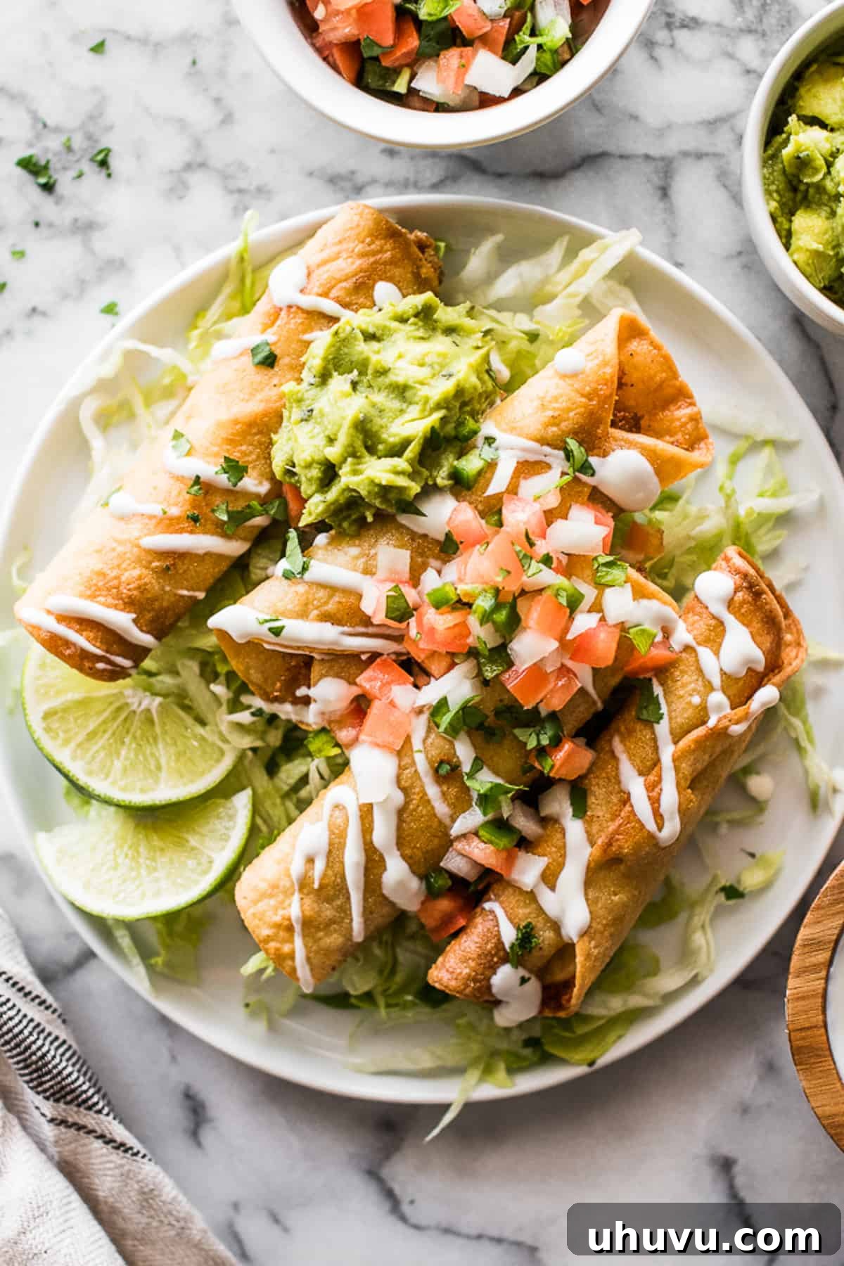 Crispy chicken taquitos on a plate garnished with Mexican crema, vibrant shredded lettuce, fresh guacamole, and zesty pico de gallo, ready to be enjoyed.