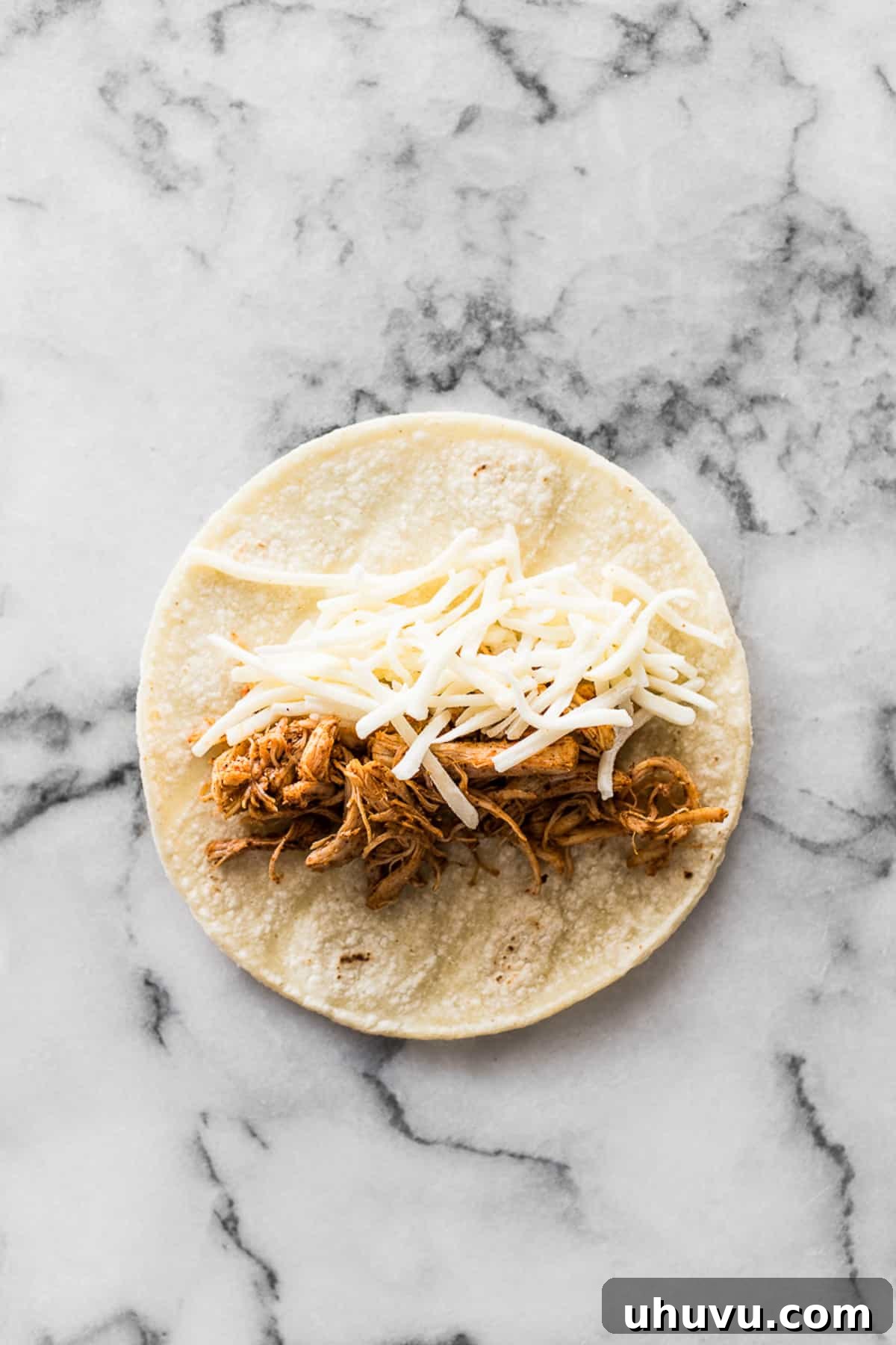 A corn tortilla with shredded cheese and seasoned shredded chicken filling placed in the middle, ready for rolling into a taquito.