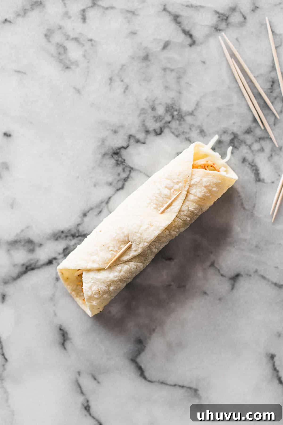 A tightly rolled corn tortilla, filled with chicken and cheese, secured shut with a toothpick, demonstrating proper taquito assembly.