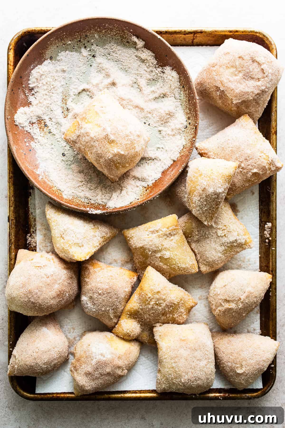 Hot sopapillas being tossed and covered in cinnamon sugar mixture in a wide, shallow dish.