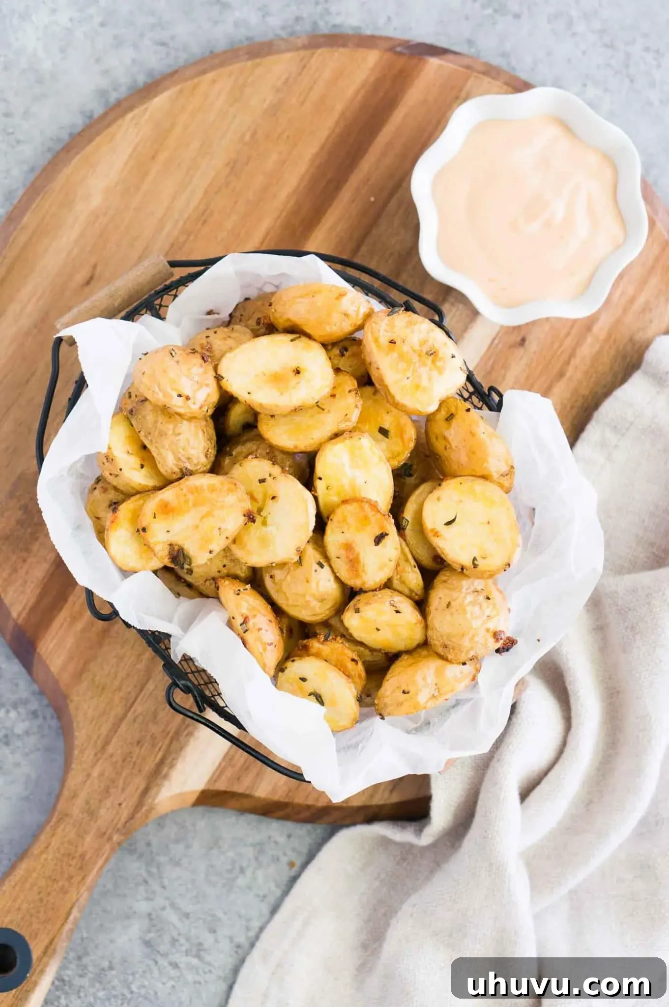 A wooden board featuring a generous serving of roasted baby potatoes alongside a bowl of dipping sauce and fresh herbs.