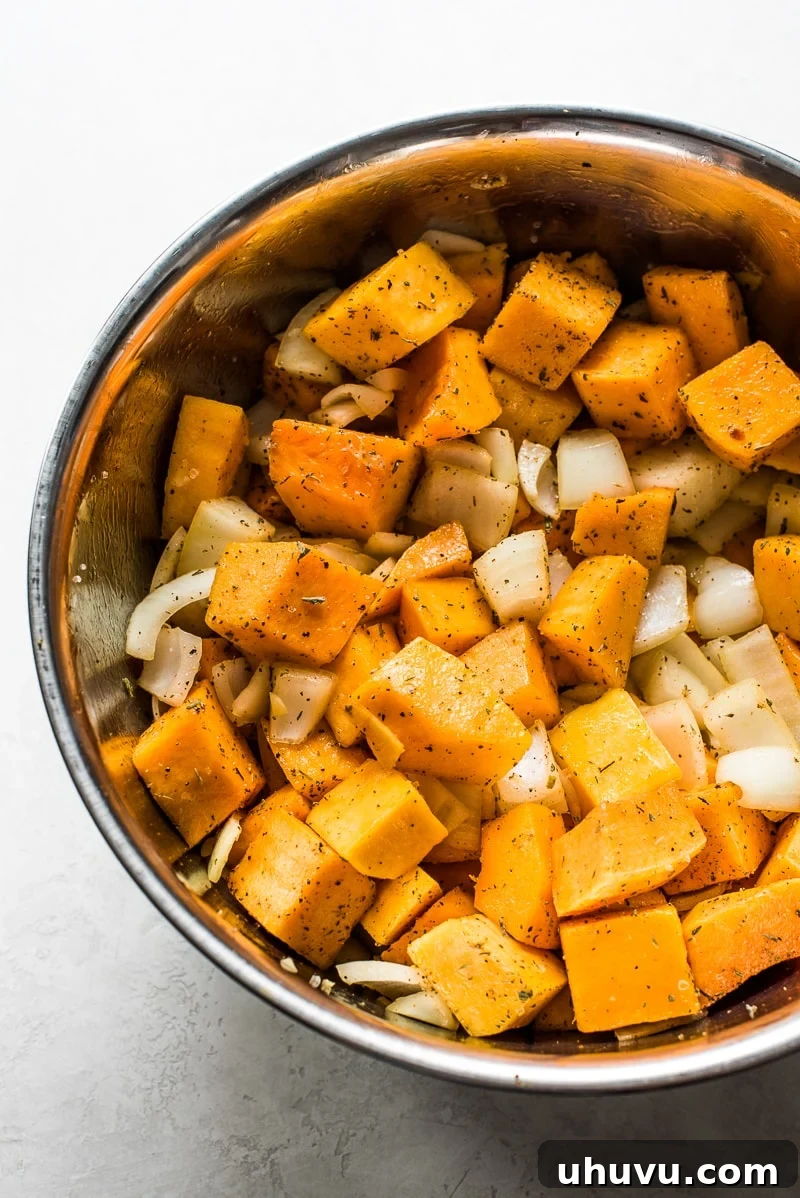 Cubed and peeled butternut squash, chopped yellow onions, and fresh herbs and spices in a large mixing bowl, ready for roasting.