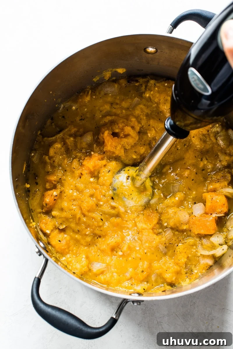Roasted butternut squash, onions, and vegetable stock being blended directly in a large stockpot using an immersion blender, creating a smooth soup base.