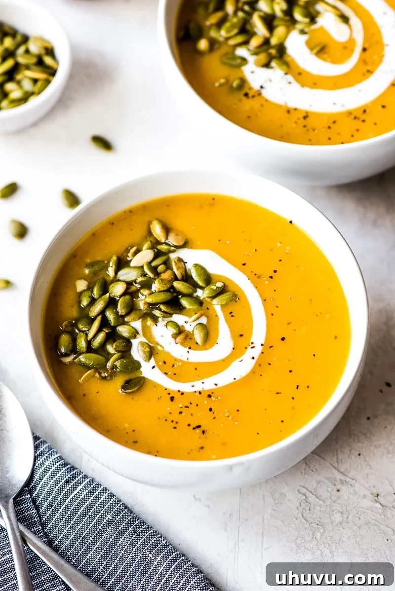 Two white bowls of creamy Roasted Butternut Squash Soup on a white wooden table, garnished with pepitas and fresh herbs.