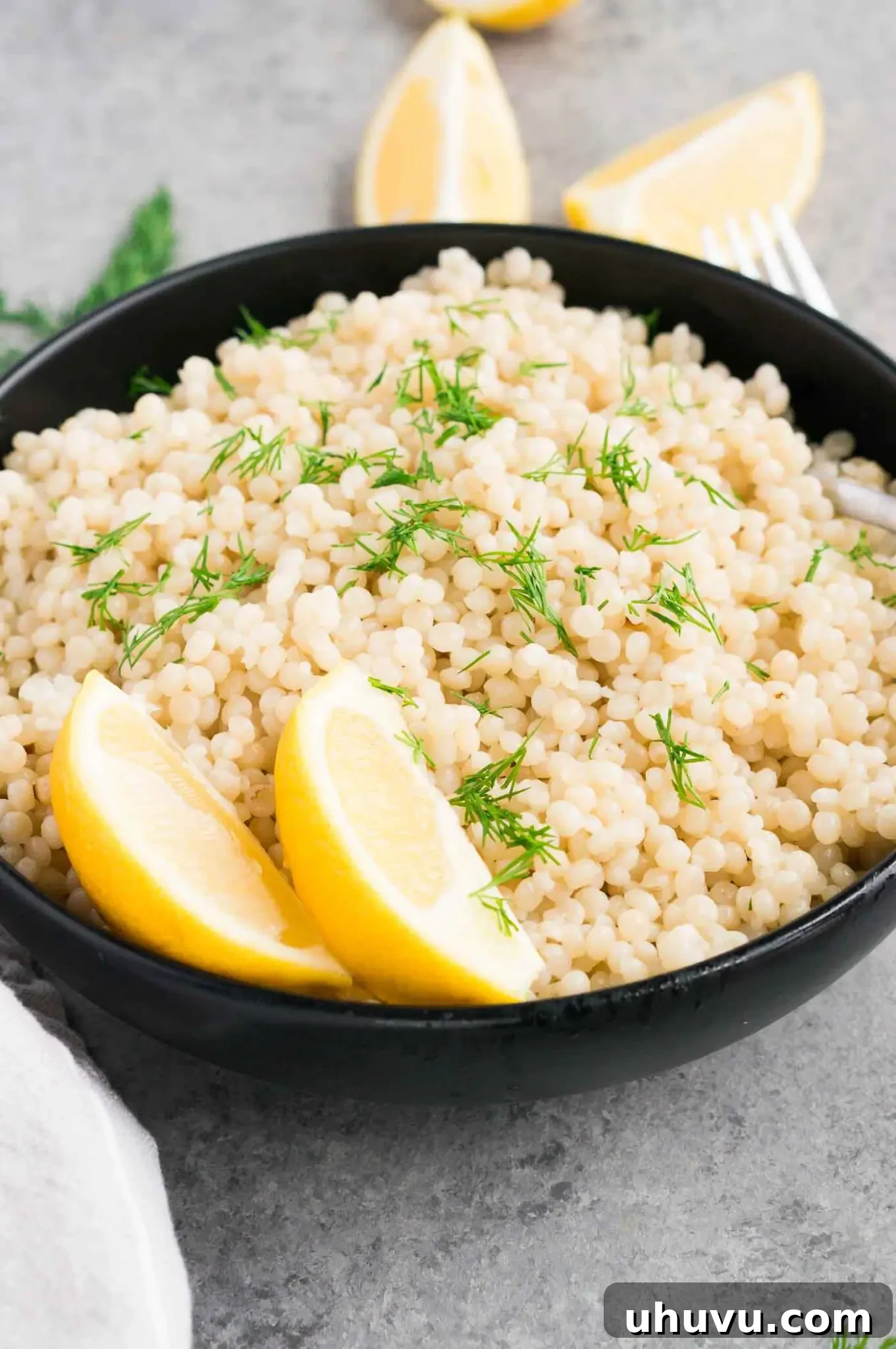 Cooked pearl couscous and lemon slices in a stylish black bowl.
