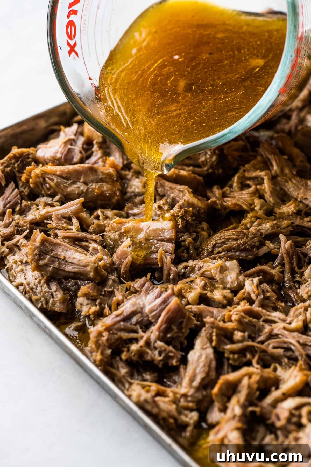 Slow-Cooked Pork Perfection 6 Flavorful cooking liquid from carnitas being drizzled over shredded pork on a baking sheet, preparing it for the crisping stage under the broiler.