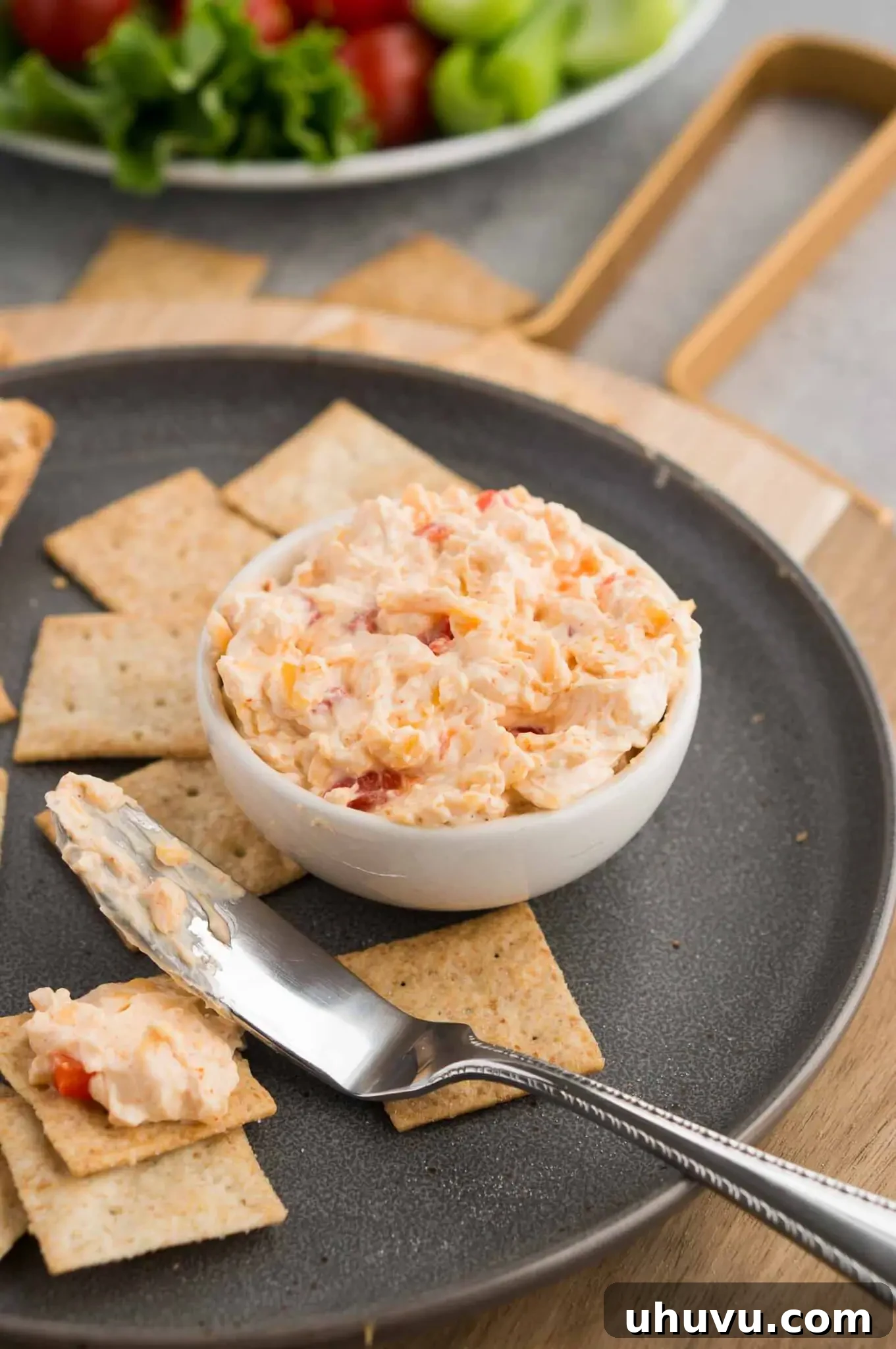 A knife spreading rich pimento cheese on a cracker, emphasizing its smooth and creamy texture.