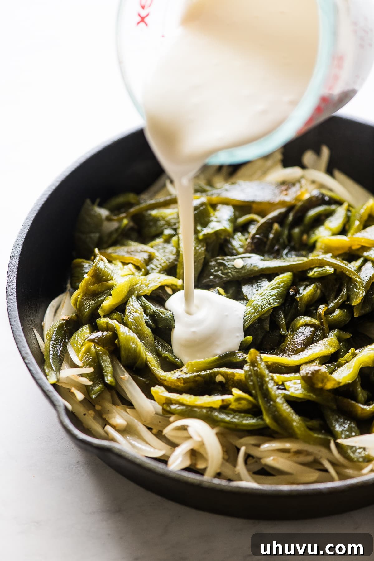 Pouring Mexican crema over sautéed poblano peppers and onions in a skillet.