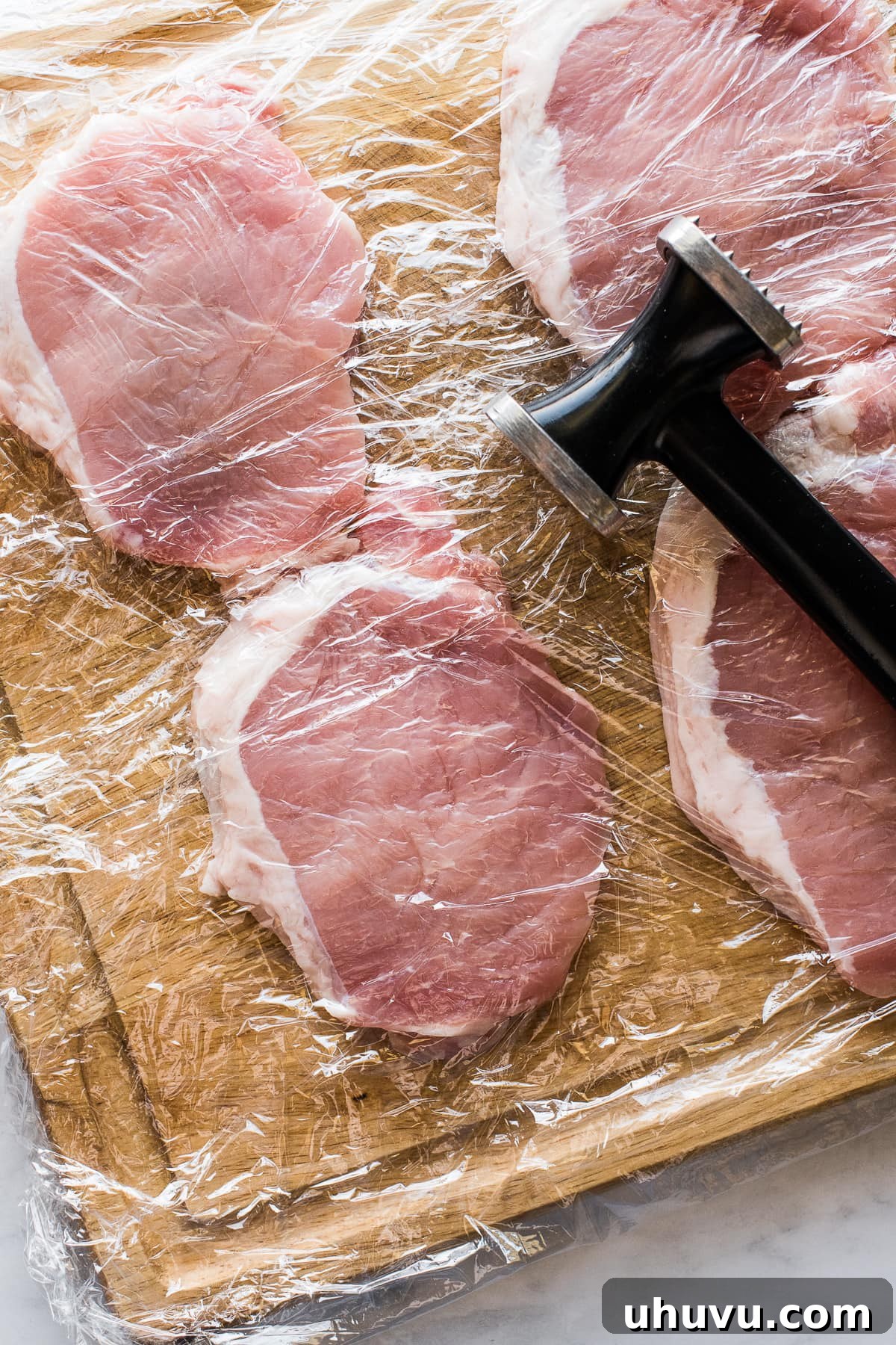 Boneless pork chops laid out on a cutting board, covered with plastic wrap, being pounded with a meat mallet.