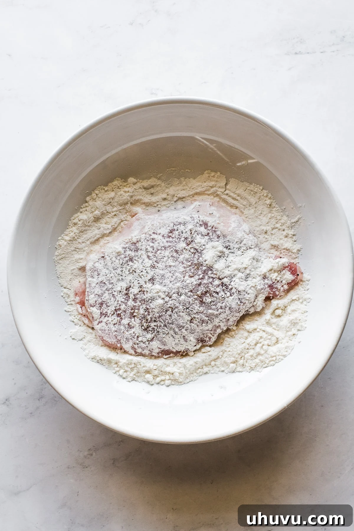 A thin pork cutlet being coated in a shallow bowl of all-purpose flour.