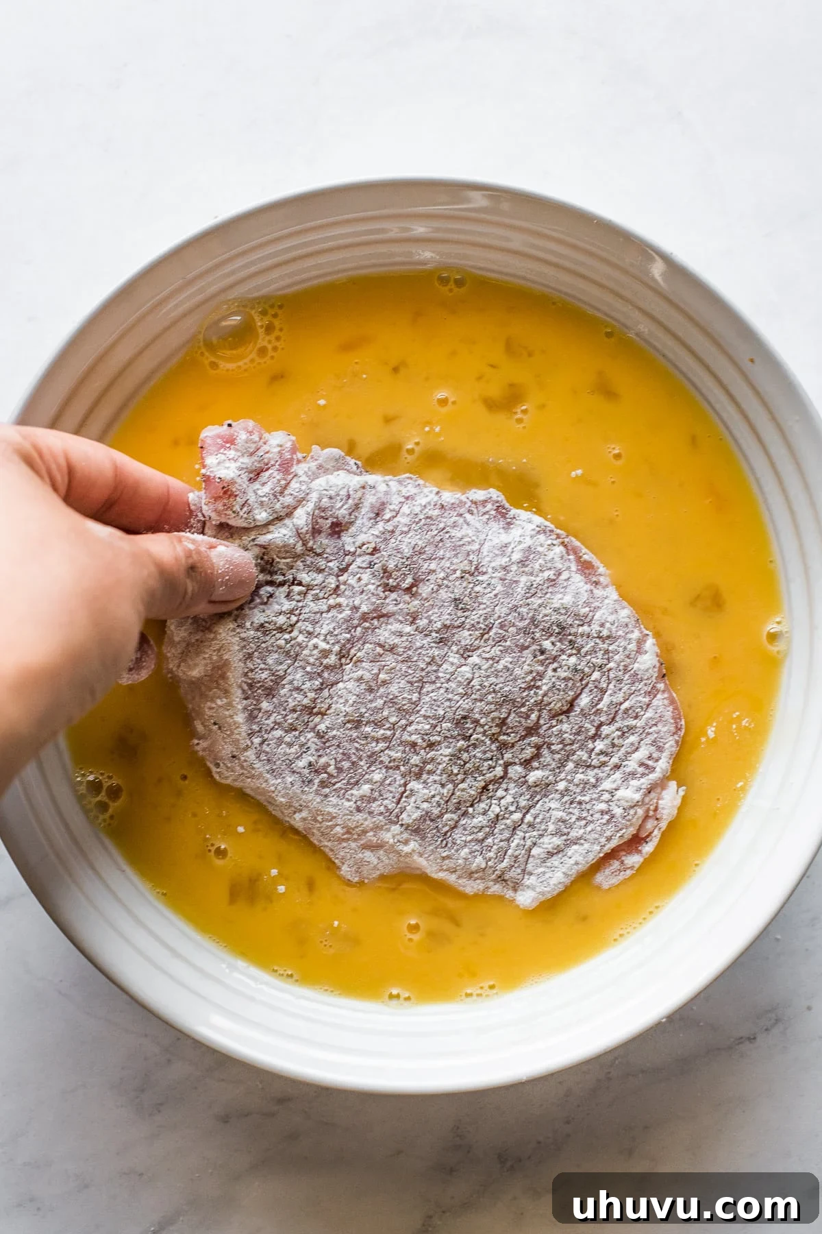 A flour-coated pork cutlet being dipped into a bowl of beaten eggs for the second stage of breading.