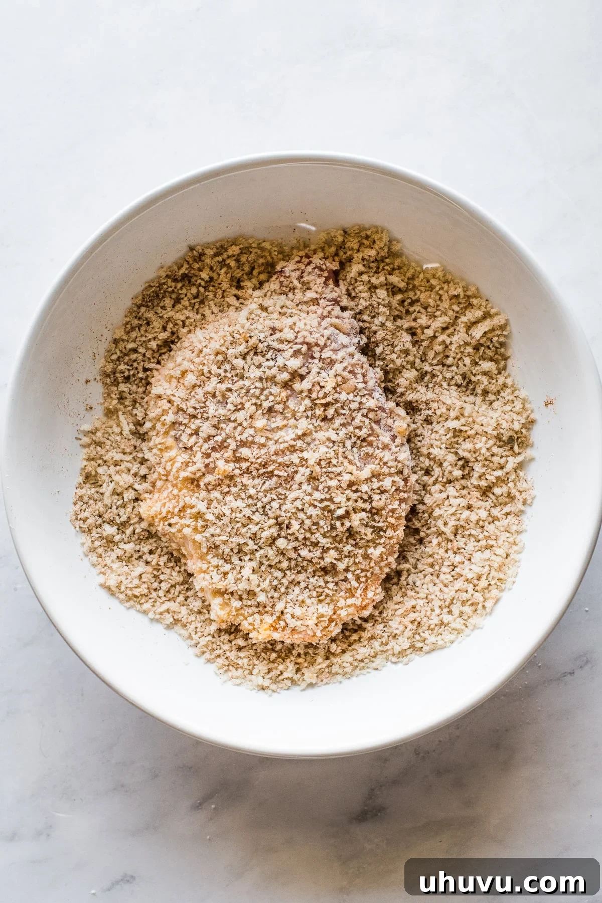 An egg-coated pork chop being pressed firmly into a bowl of seasoned panko breadcrumbs.