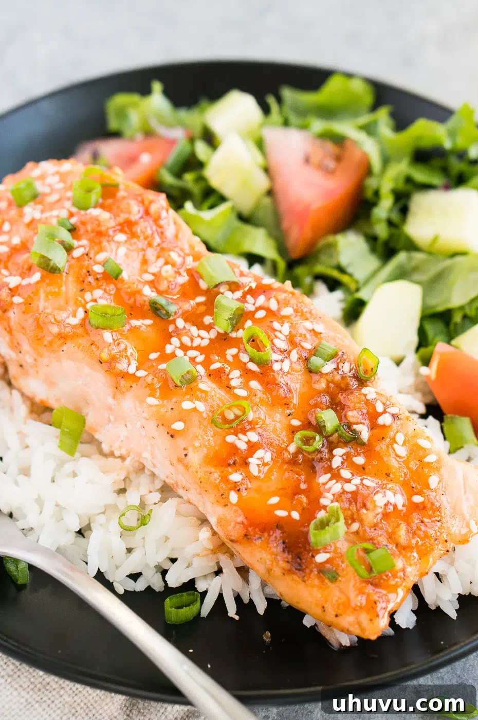 A close-up view of a perfectly cooked honey sriracha glazed salmon fillet, garnished with sesame seeds and green onions, served over a bed of fluffy white rice, ready to eat.