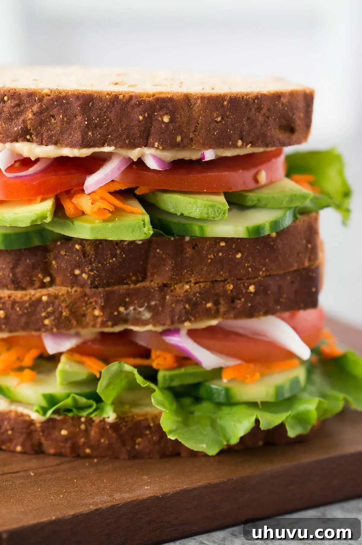 Two halves of a stacked vegetable and hummus sandwich, revealing layers of colorful fresh ingredients, presented on a wooden board.