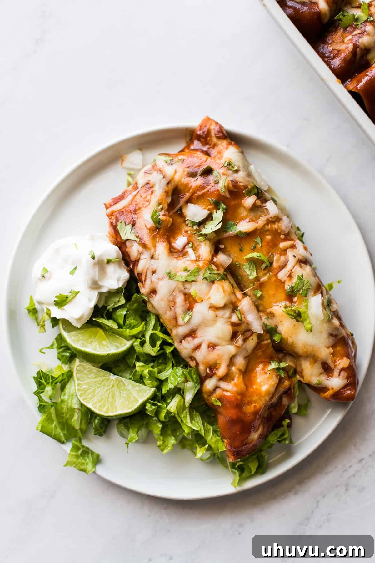 Savory Turkey Enchilada Bake 5 Two golden-brown Turkey Enchiladas served on a white plate, accompanied by fresh lime wedges, crisp lettuce, and a generous dollop of sour cream, against a rustic backdrop.
