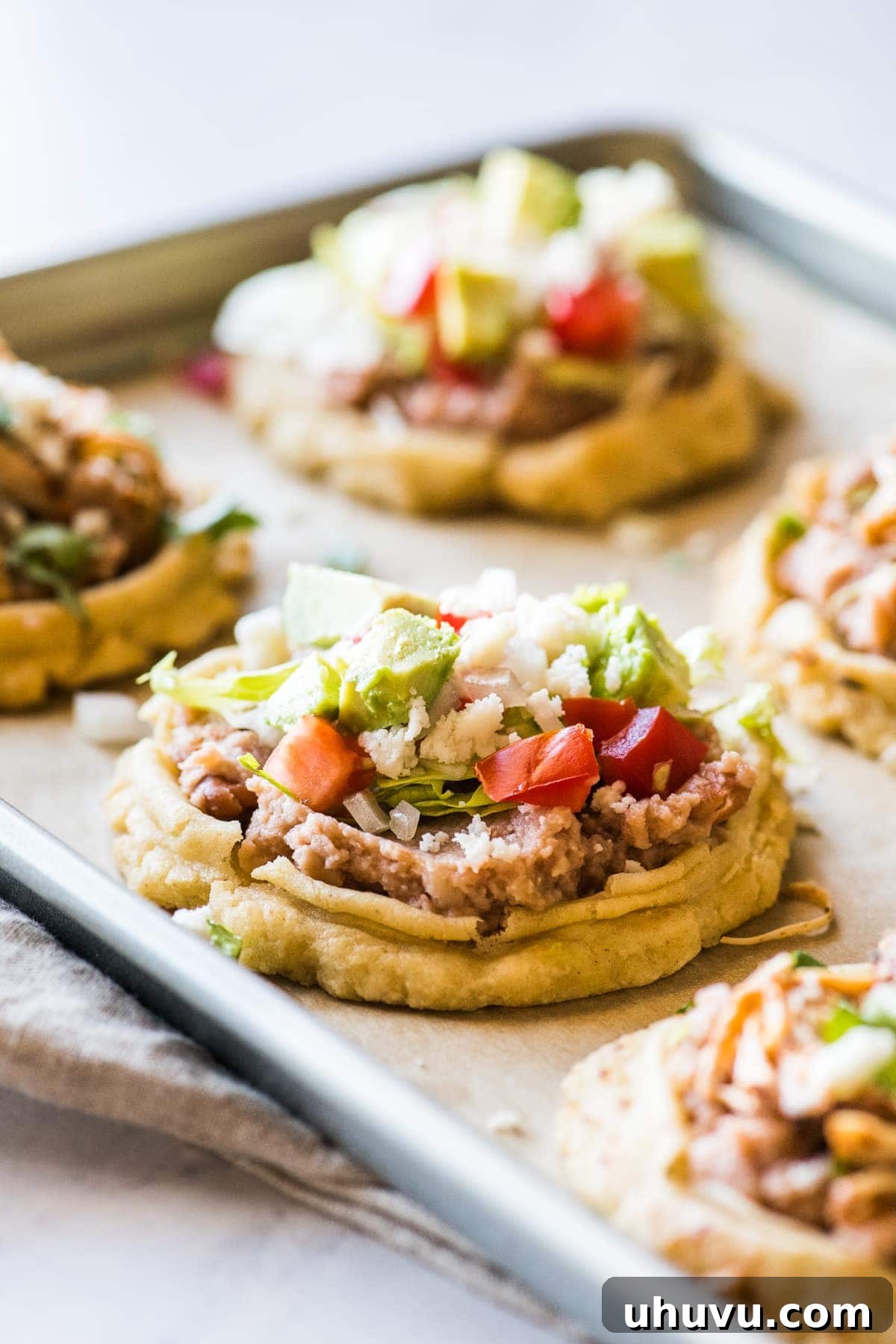 Sopes 8 Sopes on a baking sheet topped with refried beans, lettuce, and tomatoes.