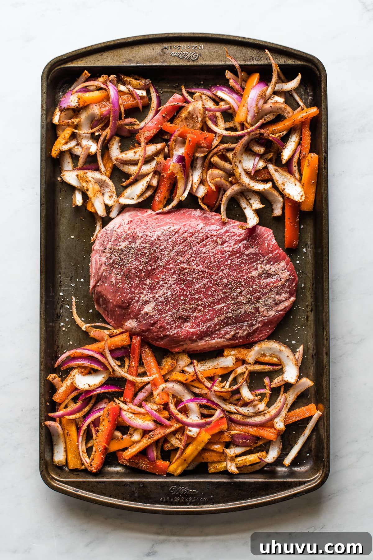 A sheet pan loaded with thinly sliced flank steak, colorful bell peppers, and red onions, all seasoned and ready for roasting.