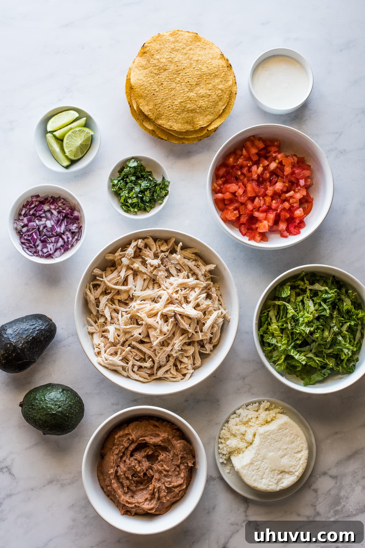 A selection of chicken tostada ingredients arranged in separate bowls, including refried beans, shredded chicken, diced tomatoes, cilantro, and cotija cheese.